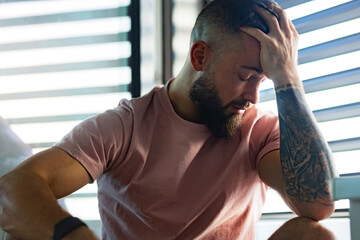 Anxious man in pink t-shirt with tattoo on arms holding head in hand.