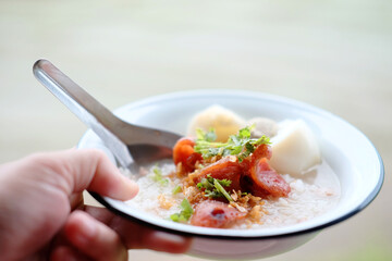 Woman hand holding rice porridge with pork and Fried Chinese sausage in white bowl.  Favorite food and tradition breakfast in Thailand