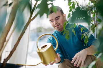 Young man with Down syndrome taking care of indoor plants, watering them, looking at camera through...