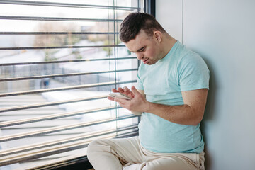 Young man with Down syndrome sitting by window, with smartphone in hand, scrolling.