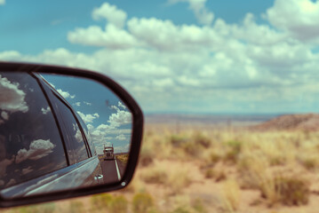 Rear view mirror of a car on a desert road, reflecting a truck in a vast desert landscape with...