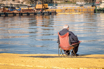 An old man sitting on a chair by the sea in the Piraeus Port of Athens Greece