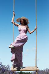 Young sexy beautiful woman swinging on hanging vintage swing in a lavender field against the background of the sky.	