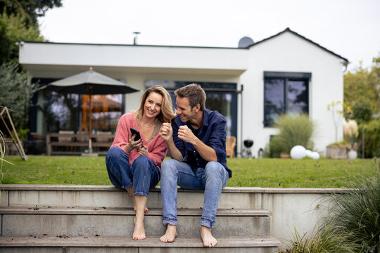 Happy couple sitting on steps with smart phone in front of house - Powered by Adobe