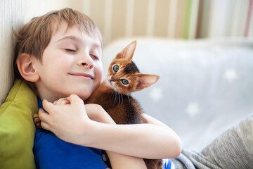 Happy child hugging his abyssinian ruddy kitten. Little boy loving red kitten at home. Pets care. ...