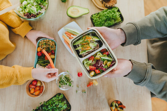 Hands of man holding vegetarian lunch box near woman at table