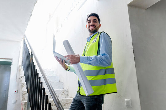 Smiling architect standing with blueprint and laptop at site