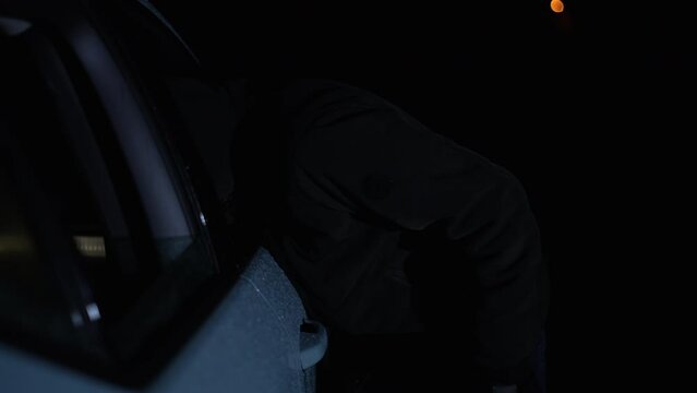 car thief smashes car window and opens the car door through the window. It is night and it is raining.