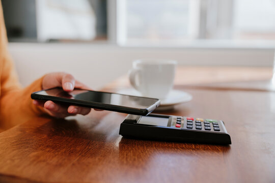 Hand of woman paying through smart phone on credit card reader near coffee cup at table