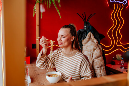 Smiling woman with eyes closed holding ramen noodles in chopsticks at restaurant