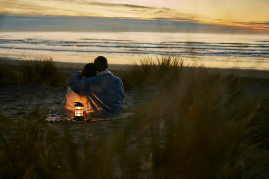 Young man with arm around of girlfriend sitting on beach at sunset