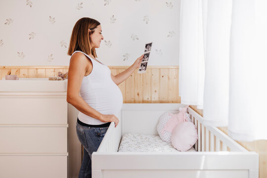 Pregnant woman holding ultrasound photograph by crib at home