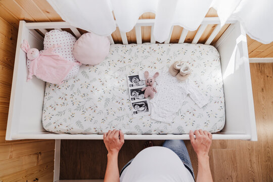 Pregnant woman standing near crib with baby ultrasound photographs and clothes at home