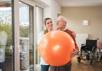 Happy home caregiver with senior man holding fitness ball and doing exercise