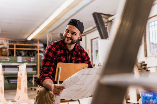 Smiling carpenter holding blueprint sitting on chair at workshop