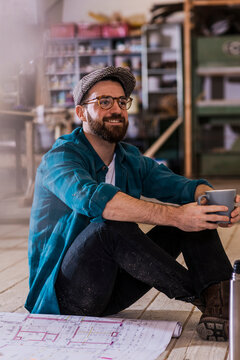 Smiling carpenter sitting by blueprint and holding coffee mug at workshop