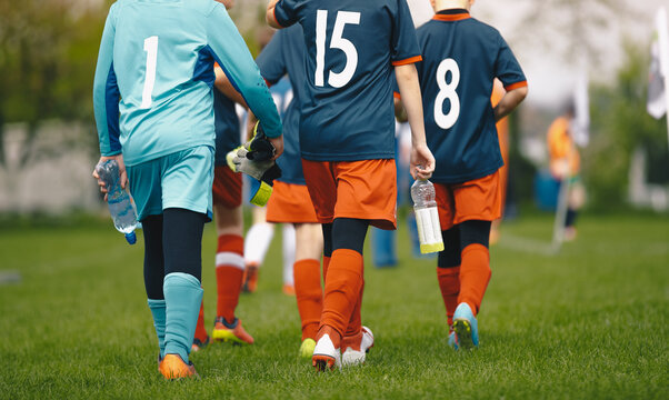 Young boys in the sports team walk off the grass pitch holding bottles of water in their hands after losing the game. Kids in blue soccer jerseys. Male elementary school sports team