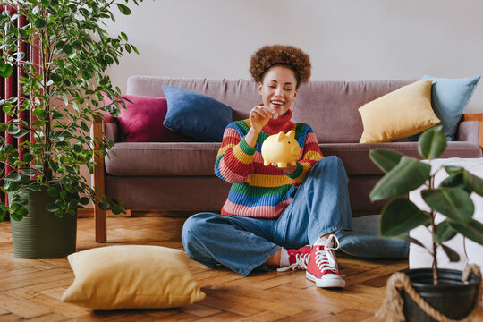 Happy young woman putting coin in piggy bank at home