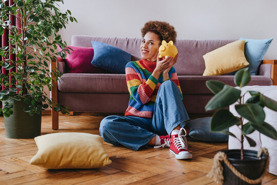 Happy young woman sitting with piggy bank at home
