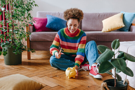 Happy young woman sitting with hammer and piggy bank at home