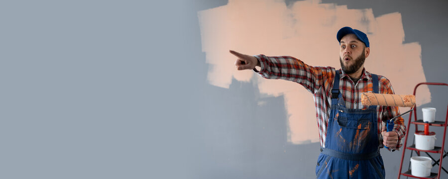 Painter man holding a paint roller surprised and pointing finger on advertising place