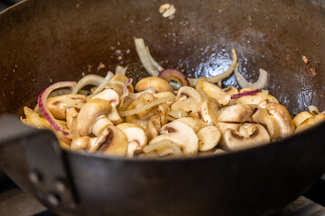 This culinary image shows sliced mushrooms and red onions being stir fried in a wok, indicative of a step in a recipe that requires sauteing vegetables to bring out their flavors. The onions are