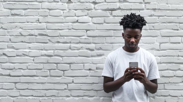 Black young man in white clothes using a smartphone against a white brick wall, fingering, reading on social networks, typing or shopping online, copy space