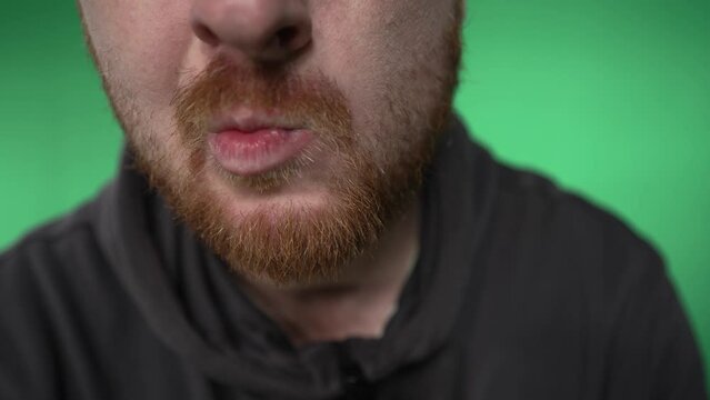 close-up of the mouth of a man with a red beard who is smiling, licking his lips and grimacing. a man with a red beard moves his lips, letting out air. male lips close up. shooting on a green back