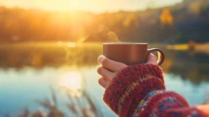 Rolgordijnen A woman wearing a sweater, holding a mug or a cup in her hands, standing outdoors, looking at the sunny lake and forest landscape in the morning. Tourist or vacation hot coffee drink, weekend beverage © Nemanja