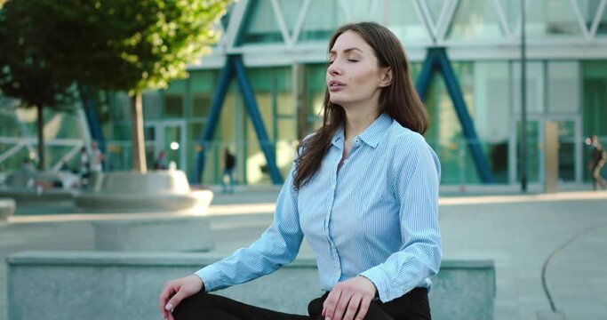 Young business woman meditates to relax her mind and body while having a break from long hard working day by sitting on bench outside office building in city center.
