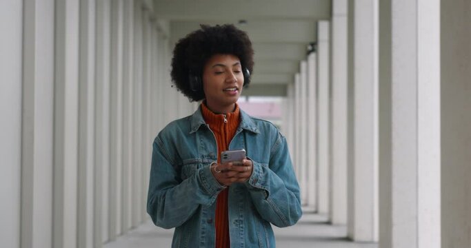 Young happy smiling african american woman with headphones is using smartphone for texting message, make calls, listen to music, browse in web and social while walking to for work or study.