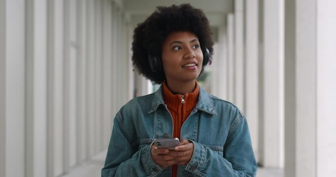 Close up of happy smiling african american woman with headphones is using smartphone for texting message, make calls, listen to music, browse in web and social while walking to for work or study.