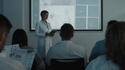 Female medical expert in white coat holding digital tablet and explaining presentation to audience while leading healthcare conference