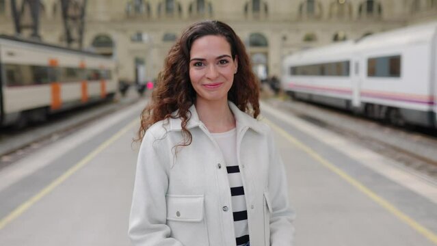 Smiling portrait of young beautiful woman in casual clothes looking at camera standing at train station platform.