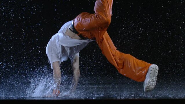 Super slow motion of young modern urban male break dancer is performing energetic street style choreography with water splashes under the rain in studio.