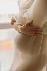 Pregnant woman in avori stylish dress standing near window and hugging yourself. Babyshower