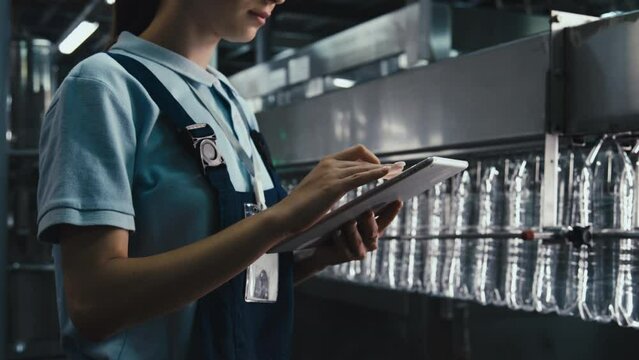 Cropped side stab footage of unrecognizable female engineer typing on tablet while standing near plastic bottles production line moving through overhead conveyor at bottled water plant