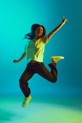 Full-length portrait of young beautiful woman jumping raising hands of joy in neon yellow lighting...