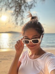 Vertical image of Asian Thai woman wear white sunglasses take selfie with sea view, while relaxing at the beach sunset time.
