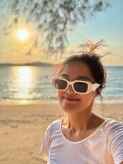 Vertical image of Asian Thai woman wear white sunglasses take selfie with sea view, while relaxing at the beach sunset time.