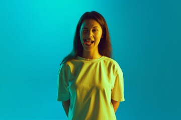 Funny young girl in white T-shirt grimacing looking at camera in yellow neon light against gradient blue studio background. Concept of human emotions, facial expression, self-expression. Ad