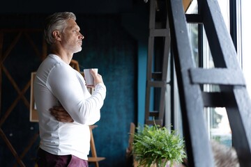 Businessman taking a coffee break standing leaning against a windowsill turning to stare out with a thoughtful expression.