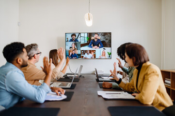 Diverse team of professionals enthusiastically participates in a video conference call, waving...