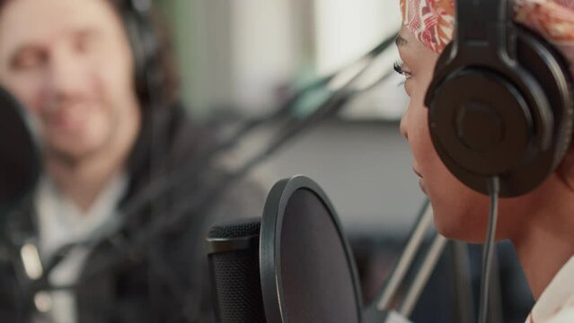 Side closeup of woman in headset talking with unidentified co-host into professional microphone during podcast in recording studio