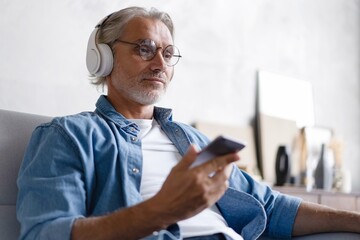 Happy Caucasian middle-aged man listening to favorite song on mobile phone with headphones on sofa...