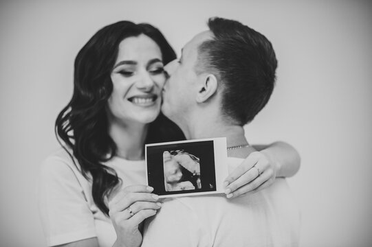Pregnant couple holding in hands ultrasound scan of their baby. Black and white photo scan. A man, husband hugging his pregnant wife in the home. Happy family pregnancy, expectation.