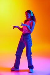 Fototapeta na wymiar Positive young lady dancing while listening music in headphones against gradient studio background in mixed neon light. Concept of human emotions, youth culture, self-expression, technology. Ad