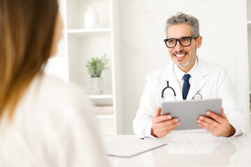 A cheerful senior doctor with grey hair and stylish glasses smiles warmly, holding a digital tablet, suggesting a modern approach to patient care. The efficiency and technological integration concept