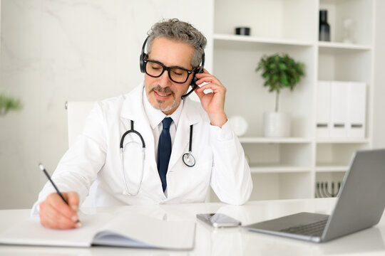 A senior grey-haired doctor in a white coat and a headset is conversing with a patient, exemplifying the personalized touch in telehealth services, attentively taking notes during virtual appointment - Powered by Adobe