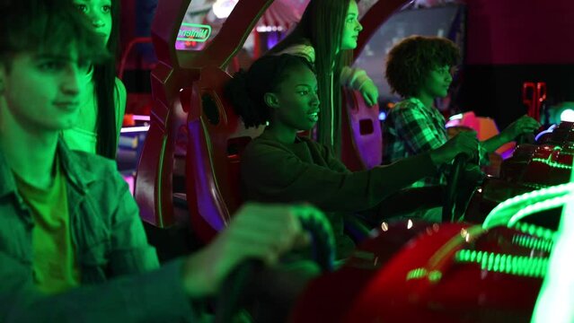 group of multiracial teenage friends in arcade playing car games with steering wheel